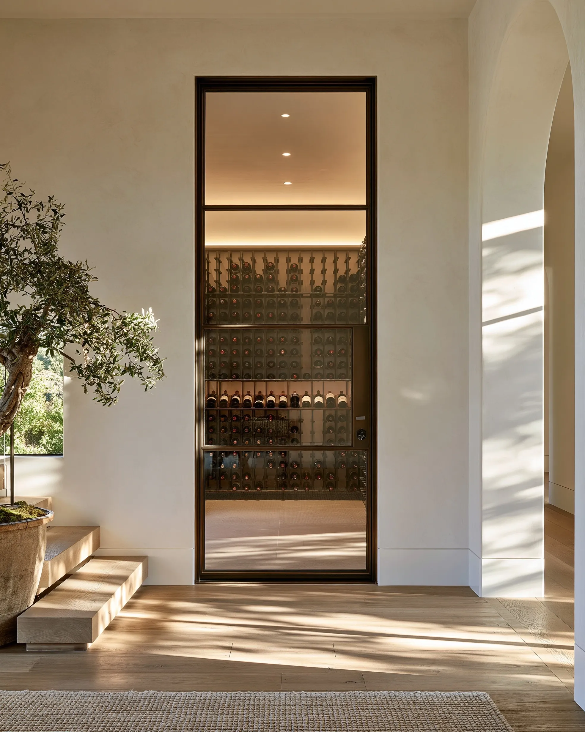 Tall dark-framed glass door into a lit wine room, set in a pale plaster hallway